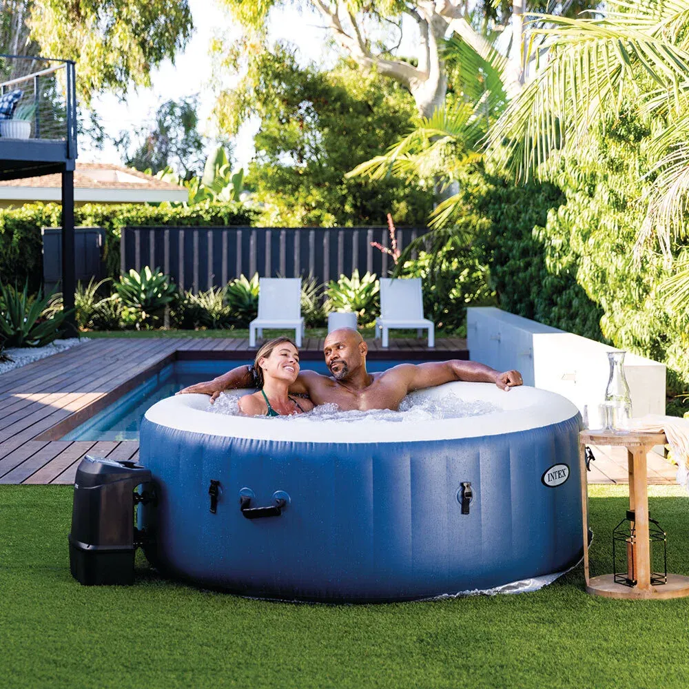 Deux adultes installés dans le spa Intex Blue One posé sur une pelouse, bloc technique à gauche