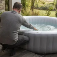 Marc, de dos, accroupi sur sa terrasse en bois, la main dans l'eau d'un spa gonflable gris, une bandelette de test posée sur le boudin, jardin au fond